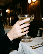 A close-up of a hand holding a coupe glass filled with sparkling white wine in a dimly lit restaurant or bar. The hand is adorned with multiple gold rings, and the background features warm candlelight and dark wood paneling.