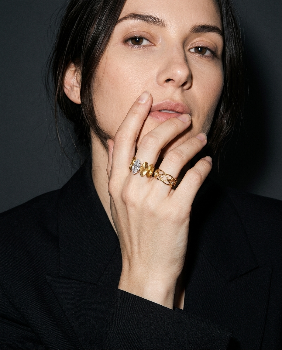 A close-up portrait of a woman wearing a black blazer and resting her hand near her face, showcasing a stack of three gold rings, including one with a clear gemstone.