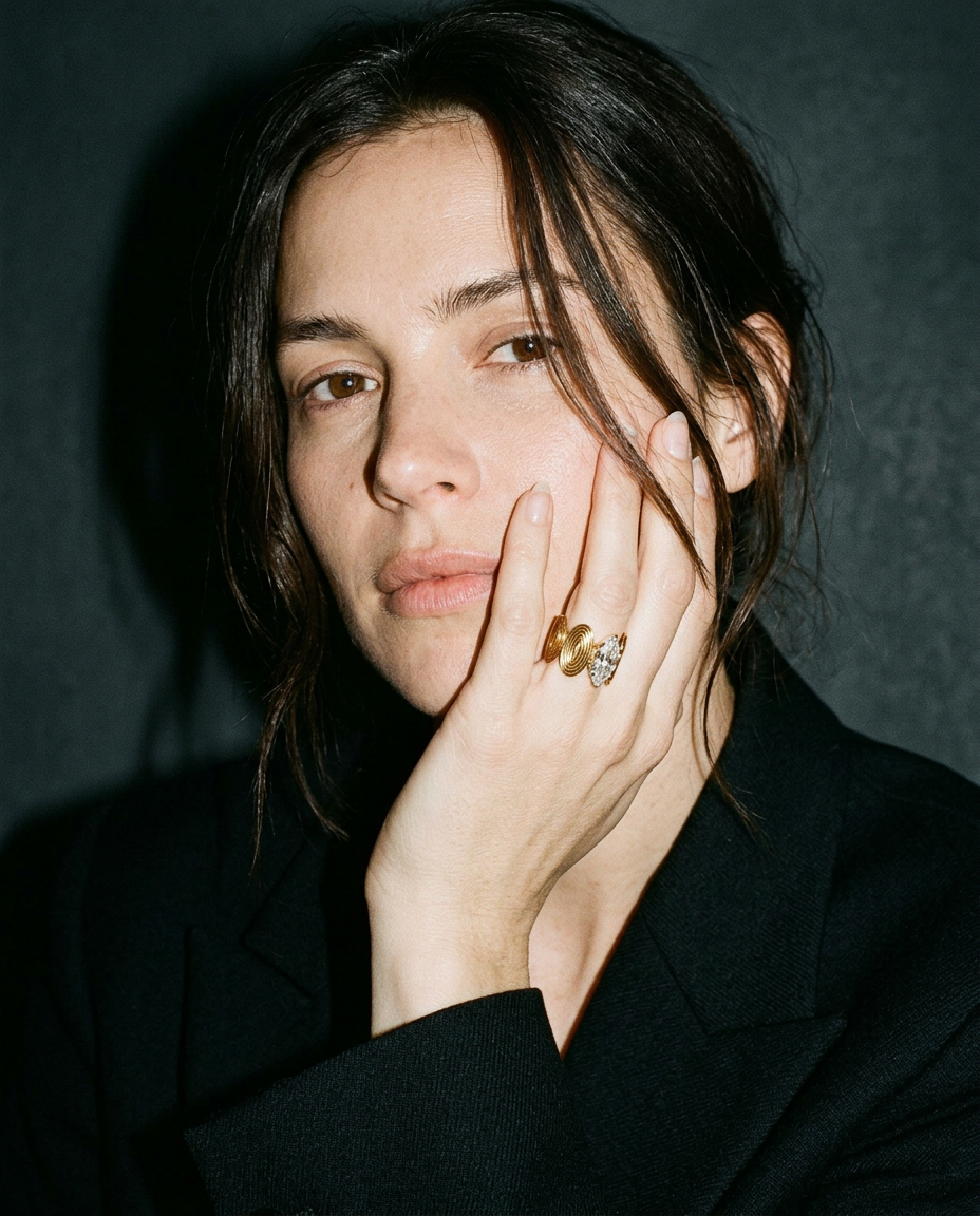 A close-up portrait of a woman with dark hair wearing a black blazer and multiple rings on her fingers, posed against a dark textured background.