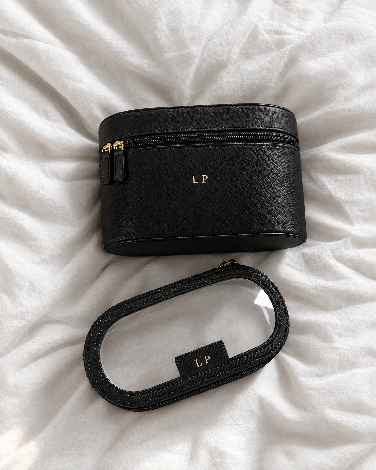 A black textured leather cosmetic bag with a matching curved strap, laid flat on white wrinkled fabric. The bag features gold hardware, a gold 'LP' monogram on the front, and a small black label on the strap with the gold 'LP' logo.