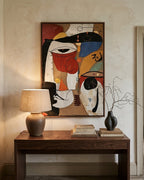 A framed abstract painting with bold geometric shapes in red, black, white, blue, yellow, and beige hangs on a textured beige wall above a dark wood console table. A lit table lamp sits on the left, and a black vase with dried branches and stacked books sit on the right.