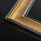 Close-up of a picture frame corner showing a distressed gold inner frame, a black outer frame, and a dark brown wood base on a black background.