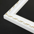 A close-up of a white decorative picture frame corner with gold embossed detailing, set against a solid black background.