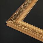 Close-up of an ornate picture frame corner with carved floral and scrollwork details in a warm gold finish, set against a dark black background.